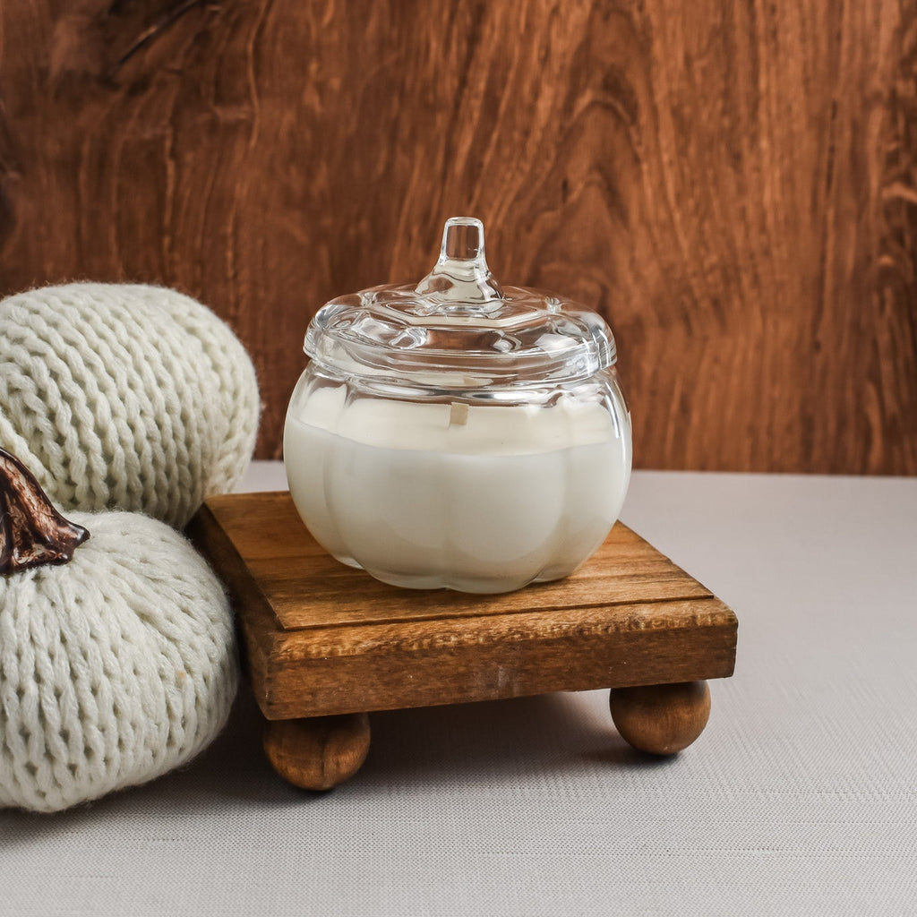 A white wax candle in a clear glass jar with a matching glass lid resting beside it on a small wooden stand. The candle is styled with two knitted cream pumpkins against a rustic dark wood background.