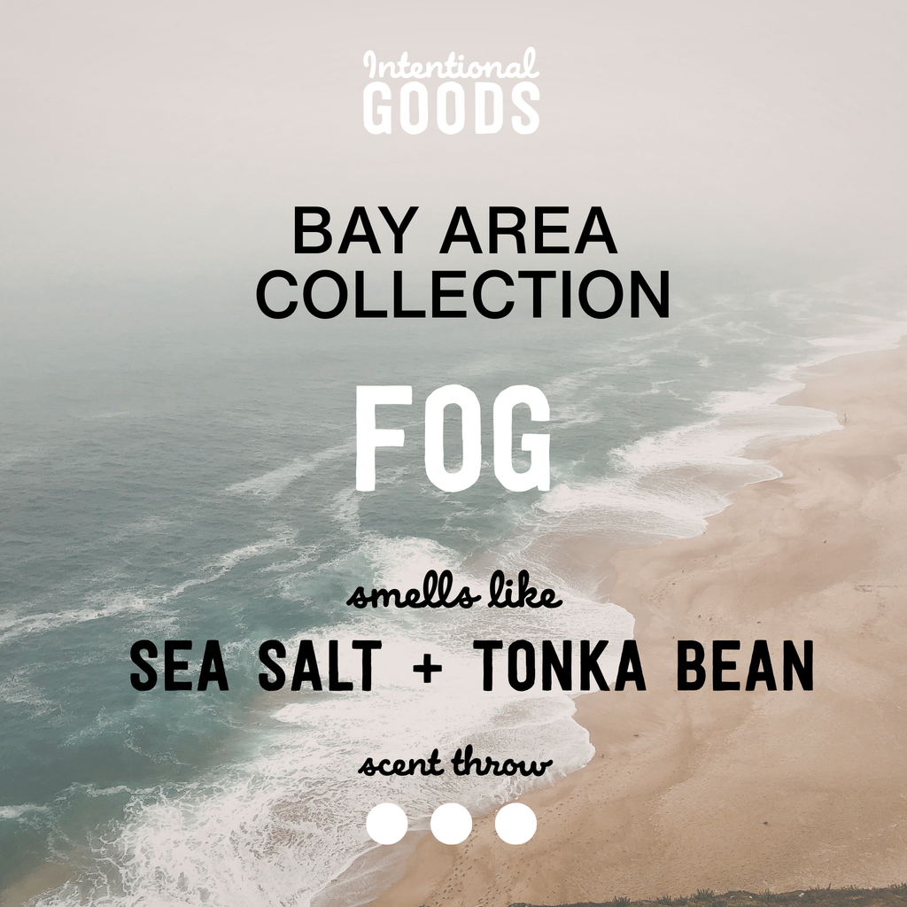 A white candle with a wooden lid labeled 'FOG' on a wooden stool, alongside a gold wick trimmer and other home décor items.