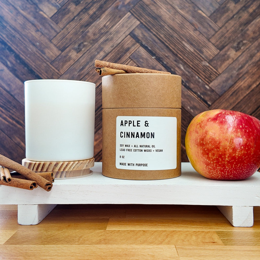 Candle in a brown paper sleeve labeled 'Apple & Cinnamon' on a woven basket with an apple.